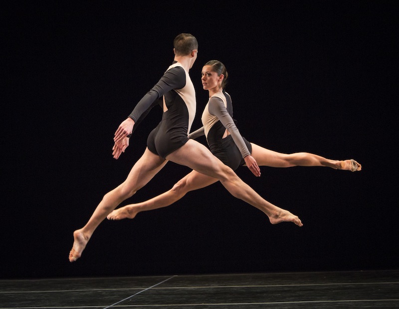 Joshua Tuason and Melissa Toogood in Locomotor.  Both dancers look at one another as they leap in the air, splitting both legs perpendicular to the floor.